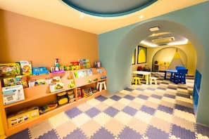 Een kinderspeelruimte met een geruit vloerkleed, planken vol speelgoed en een tafel met stoelen.