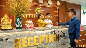 A reception desk with a marble top and the word "RECEPTION" prominently displayed. There are clocks above the desk indicating different time zones, and a decorative vase with dried plants. - Aloha Hotel (Hanoi)
