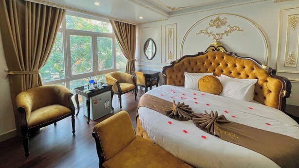 A luxurious hotel room with a large bed, two armchairs, a small table, and a view of greenery through the window. - Aloha Hotel (Hanoi)
