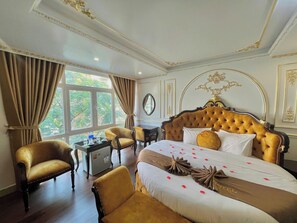 A luxurious hotel room with a large bed, two armchairs, a small table, and a view of greenery through the window. - Aloha Hotel (Hanoi)