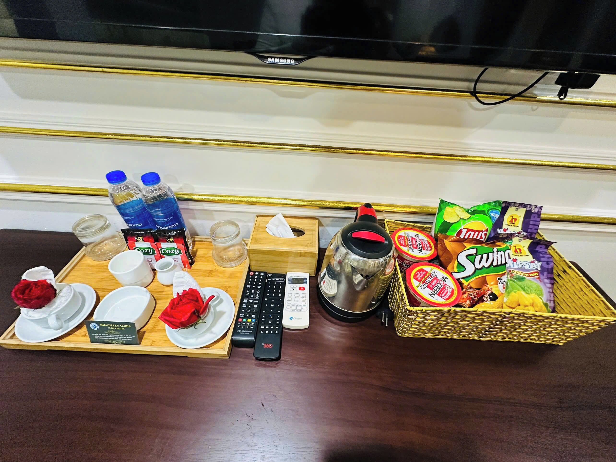 A tray with a remote control, a kettle, snacks, and a bottle of water on a wooden table.