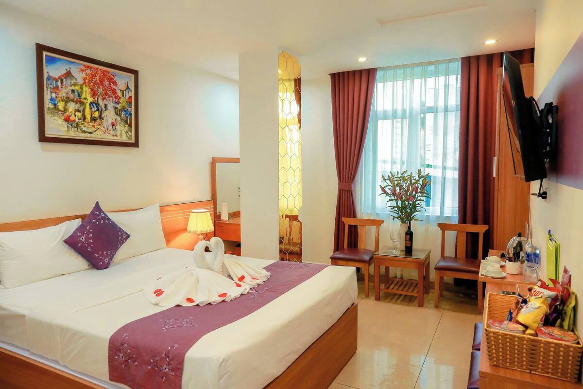 Una habitación de hotel con cama, un cuadro, televisor, ventana con cortinas, una mesita, una silla y una planta.