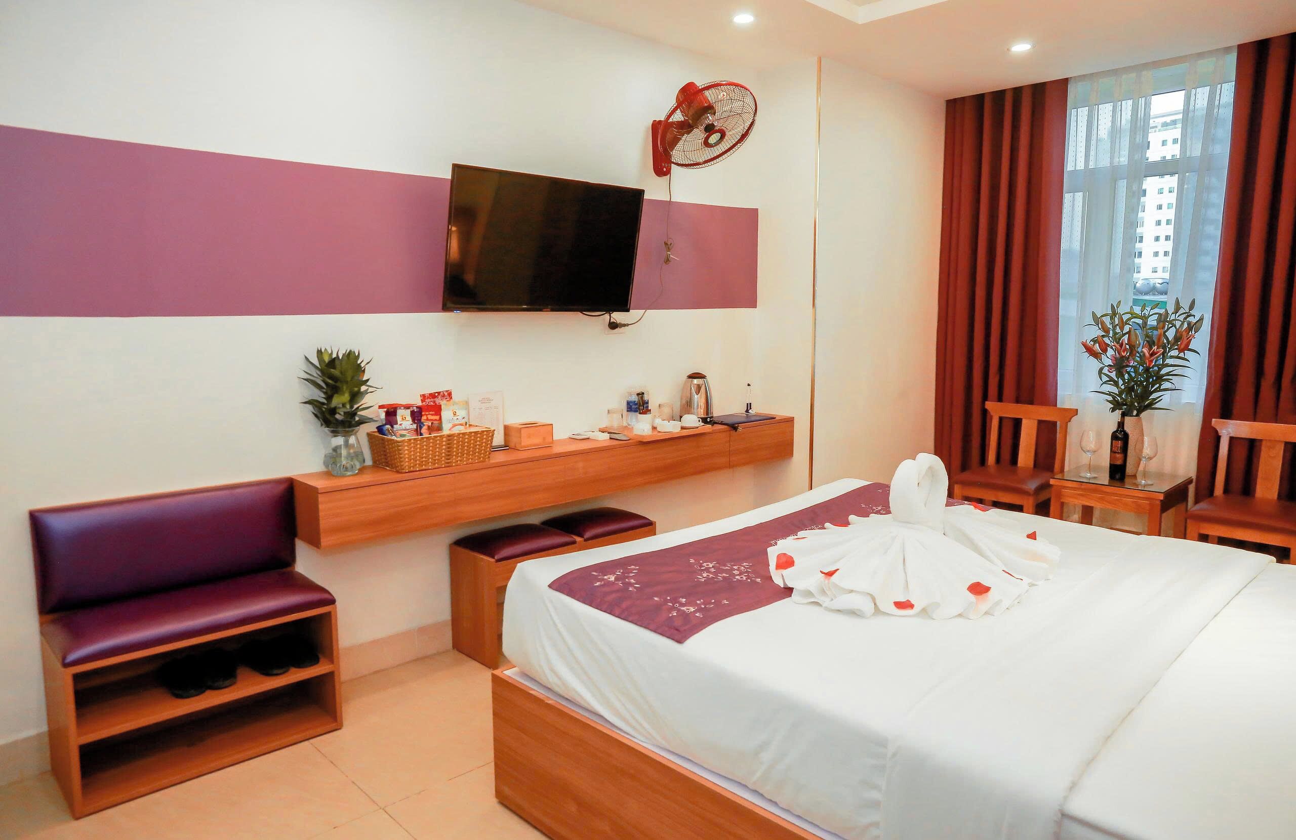 A hotel room with a bed, a TV mounted on the wall, a small seating area, a bedside table with a lamp, and a window with curtains.