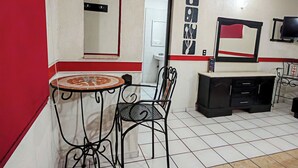 Une chambre d’hôtel de taille modeste, comprenant une petite table à manger et une chaise, une chaise en métal noir, une commode noire et un téléviseur posé sur un meuble.