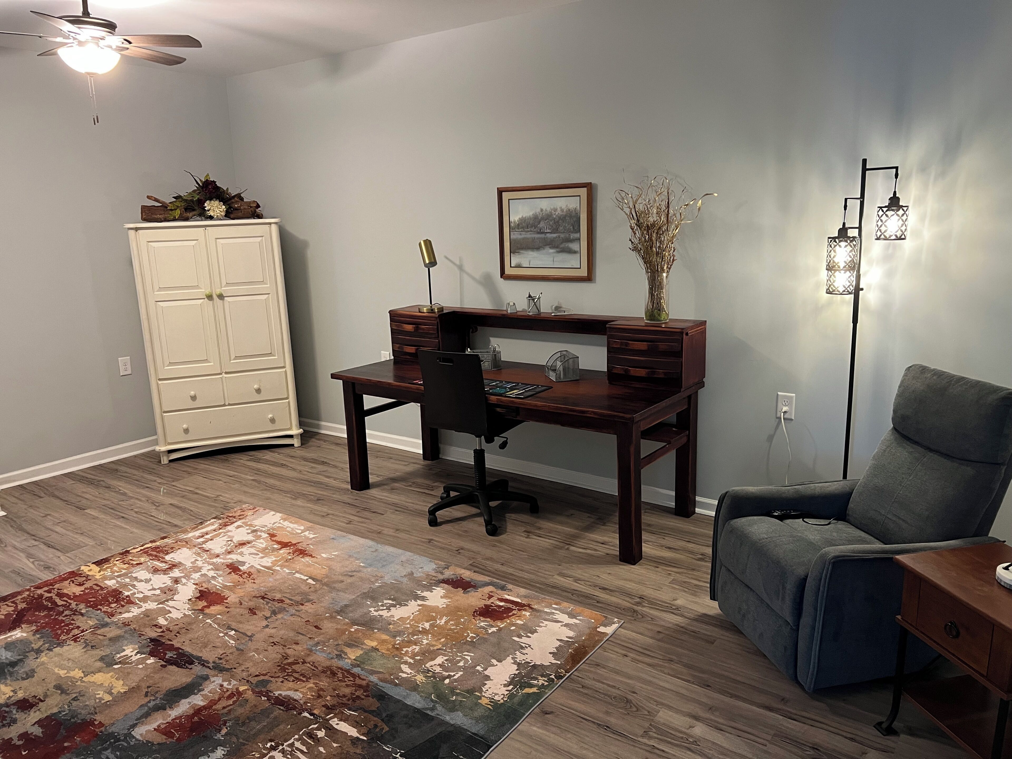 A room with a desk, chair, wardrobe, rug, and a ceiling fan.