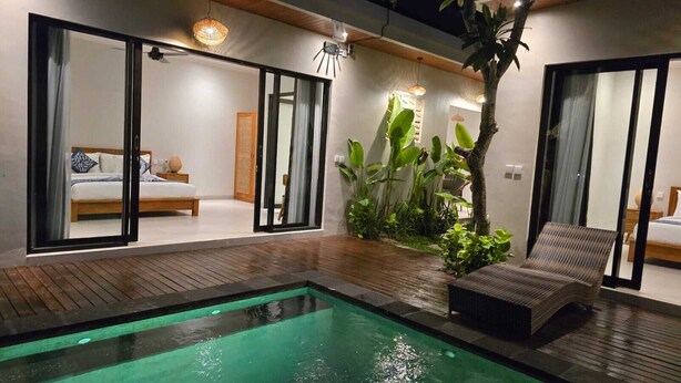 A modern indoor pool area with a wooden deck, a lounge chair, and a bed visible through a glass door. - Adyatma Ubud (Ubud)
