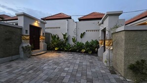 A courtyard with a paved walkway, a dark entrance door, and a white wall with a red-tiled roof. - Adyatma Ubud (Ubud)