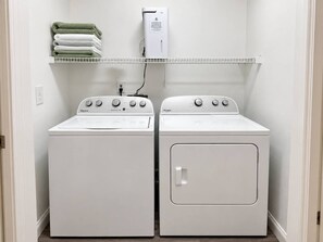 A laundry room with two Whirlpool washers and dryers, a white shelf with folded towels, and a power outlet. - Modern Top-Floor Loft w/ Pool, Balcony & Garden View! (Wilmington)