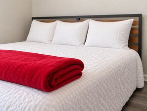 Un lit bien fait, avec une couette blanche et un plaid rouge, une tête de lit en bois et un mur sobre.