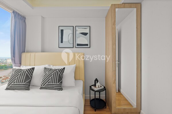 Une chambre d’hôtel moderne avec un lit, deux œuvres d’art abstraites encadrées, une petite table d’appoint avec un livre et une bouteille, et une vue sur la ville par la fenêtre.