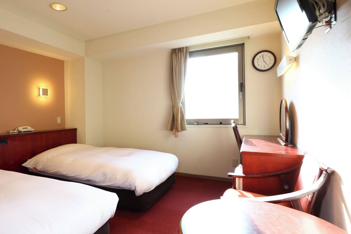 Ein Hotelzimmer mit zwei Einzelbetten, einem Schreibtisch mit Stuhl, einer Wanduhr und einem Fenster mit Vorhängen.