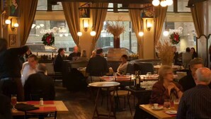 Interior de restaurante con varias mesas y sillas, iluminación cálida y ventanas amplias con cortinas.