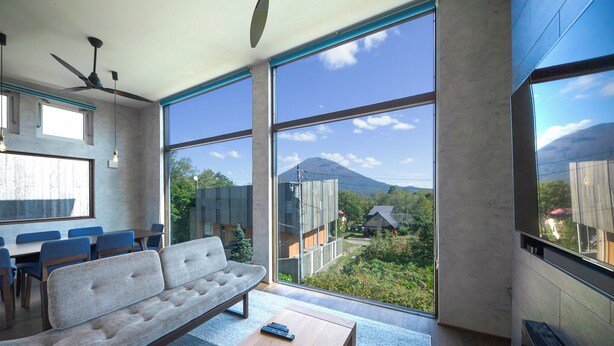 A modern living room with a sofa, coffee table, and large windows offering a view of a mountain and surrounding greenery. - Off Piste Chalet (Kutchan)