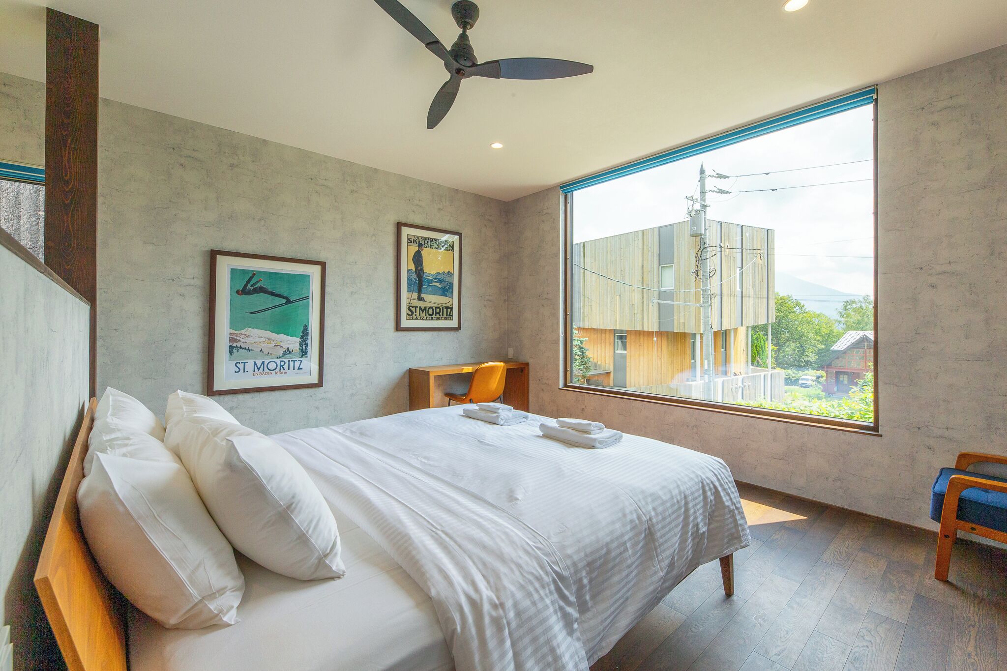 A modern hotel room with a large bed, a desk, and a chair. There are two framed pictures on the wall, a ceiling fan, and a large window with a view of buildings and greenery.