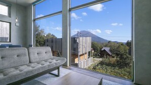 A modern living room with a large window, a sofa, and a view of a mountain. - Off Piste Chalet (Kutchan)