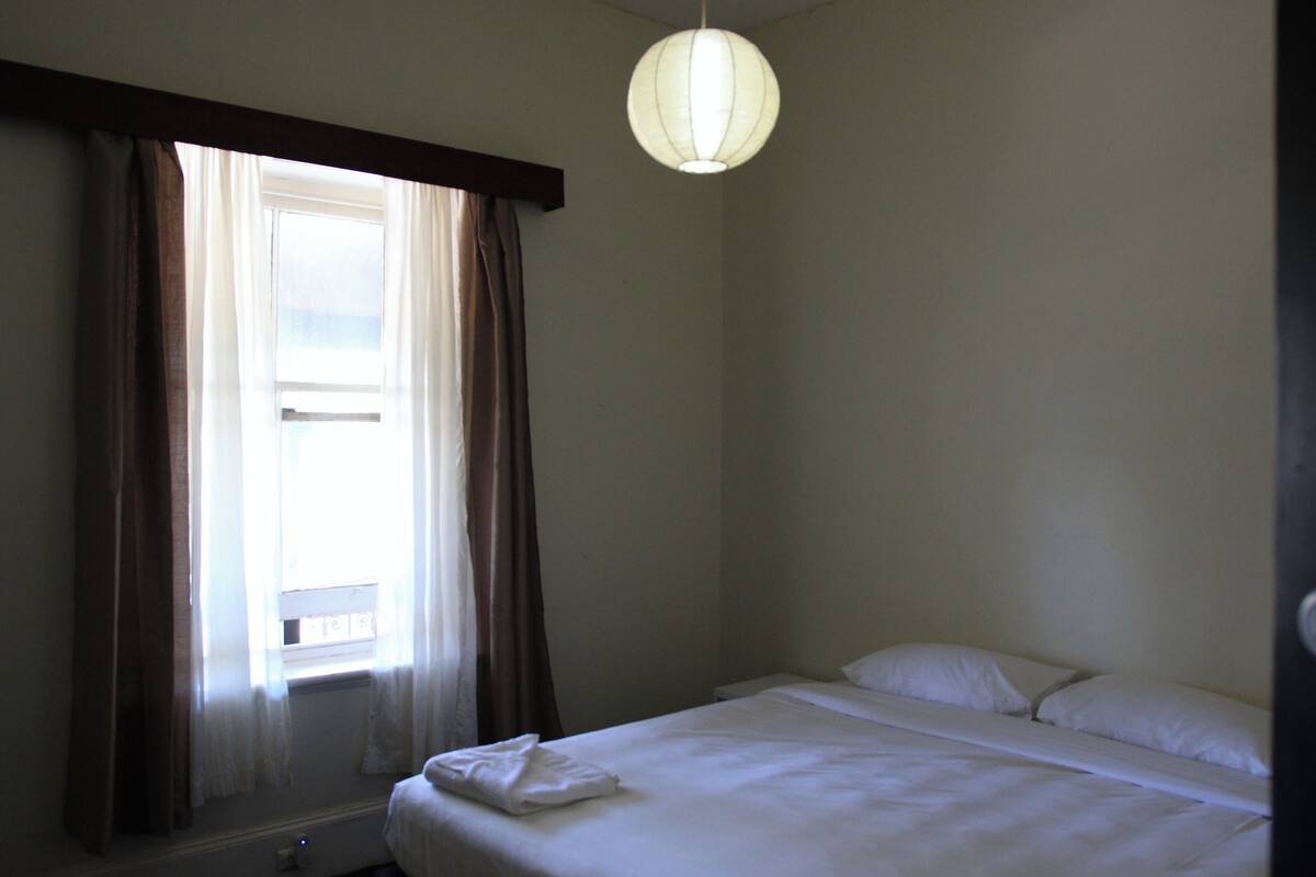 Ένα δωμάτιο ξενοδοχείου με ένα μονό κρεβάτι, ένα παράθυρο με κουρτίνες και ένα φωτιστικό που κρέμεται από την οροφή.