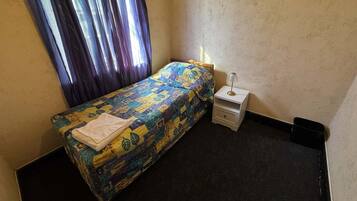 Una habitación pequeña con cama individual, colcha estampada, mesita de noche con lámpara y ventana con cortinas.