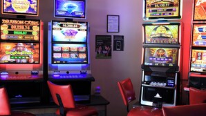 Eine Reihe von Spielautomaten mit verschiedenen Spielmotiven und ein kleiner Tisch mit Stühlen in einem schwach beleuchteten Raum.