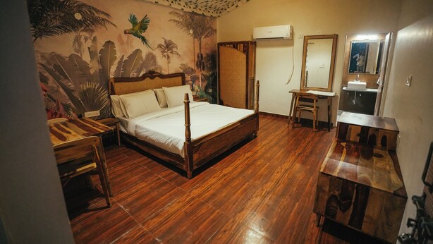A room with a bed, a wooden desk, a mirror, and a bathroom with a sink and mirror. - Vanantara Courtyard (Alwar)