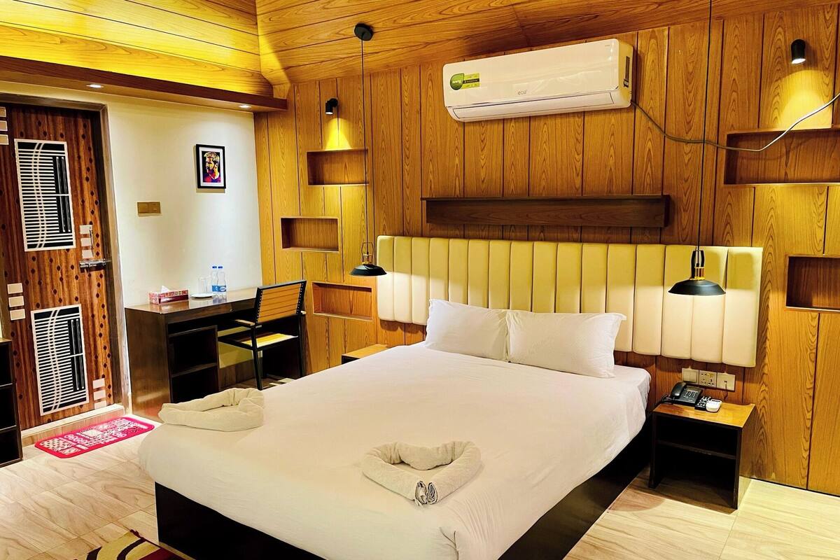 A hotel room with a wooden paneled wall, a bed with white linens, two bedside tables with lamps, and an air conditioning unit.