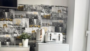 Uma cozinha compacta com geladeira branca, uma mesa pequena e uma torradeira. Na parede, uma colagem de fotografias em preto e branco de paisagens urbanas.
