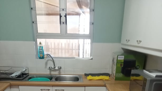 Une cuisine avec des meubles blancs, un évier, une cuisinière et une fenêtre avec des stores.