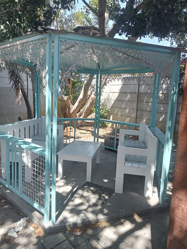 A gazebo with white benches and a metal gate, surrounded by trees and a concrete wall.