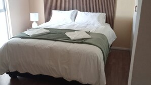 Un lit bien fait, avec du linge de lit blanc et un plaid vert, une lampe de chevet et une tête de lit en bois.