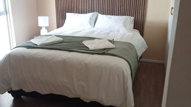 Una cama bien hecha con sábanas blancas y un plaid verde, una lámpara de noche y un cabecero de madera.