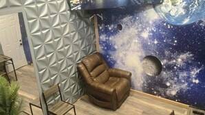 Ein Zimmer mit Wandgemälde von Stadtansichten, ein brauner Ledersessel, ein kleiner Tisch und eine Topfpflanze.