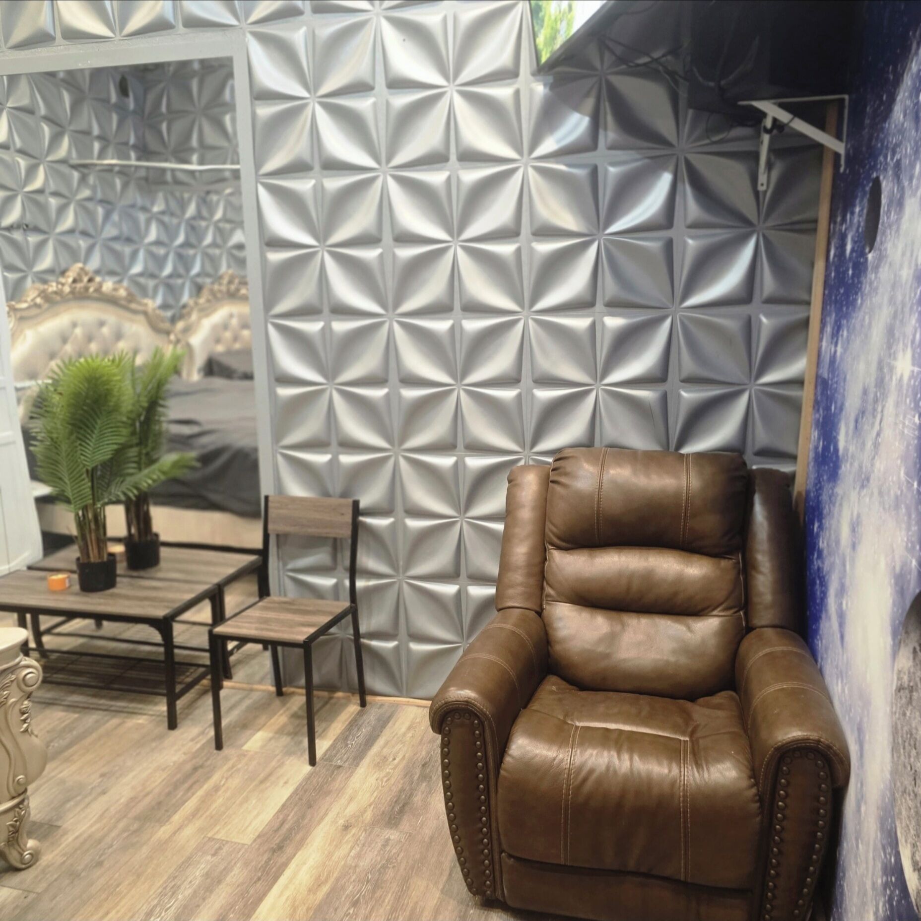 Un fauteuil en cuir marron dans une pièce dont le mur est orné d’un motif géométrique et qui contient un lit avec une tête de lit capitonnée.