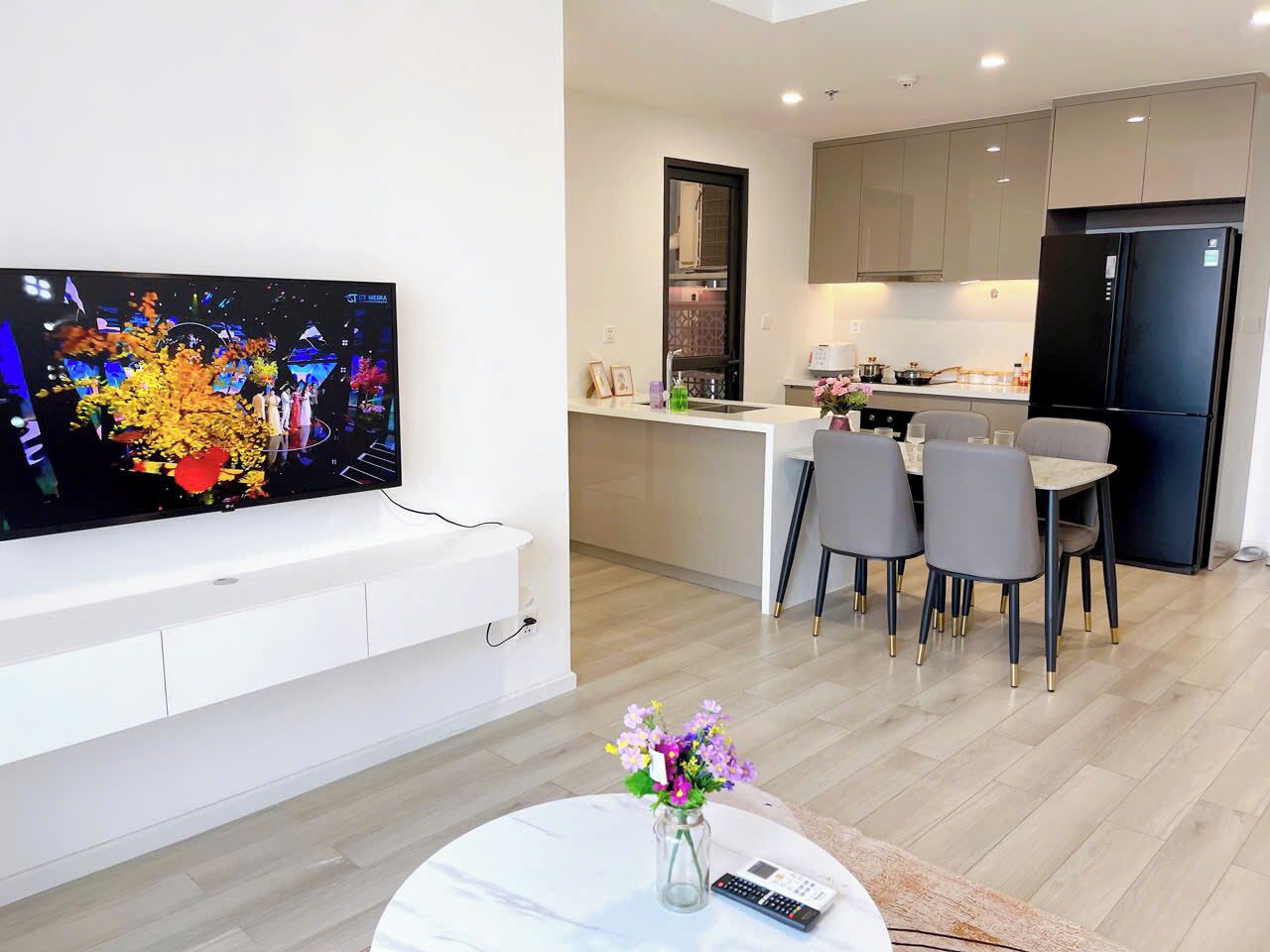 A modern living room with a flat-screen TV, a dining area with chairs, a kitchen with a refrigerator, and a small table with a vase of flowers.