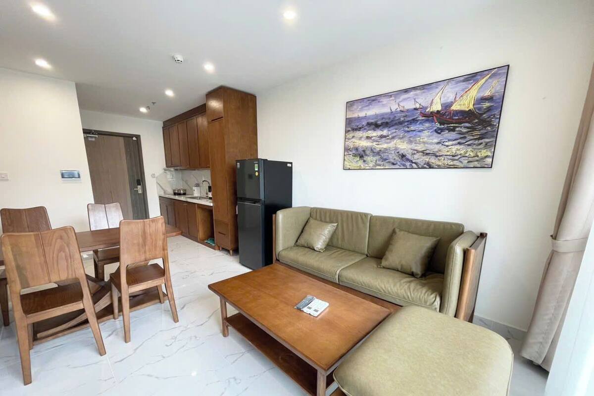 A modern living room with a wooden dining table, beige sofa, and a coffee table. There is a kitchen area with wooden cabinets and a refrigerator. A painting of a sailboat is on the wall.