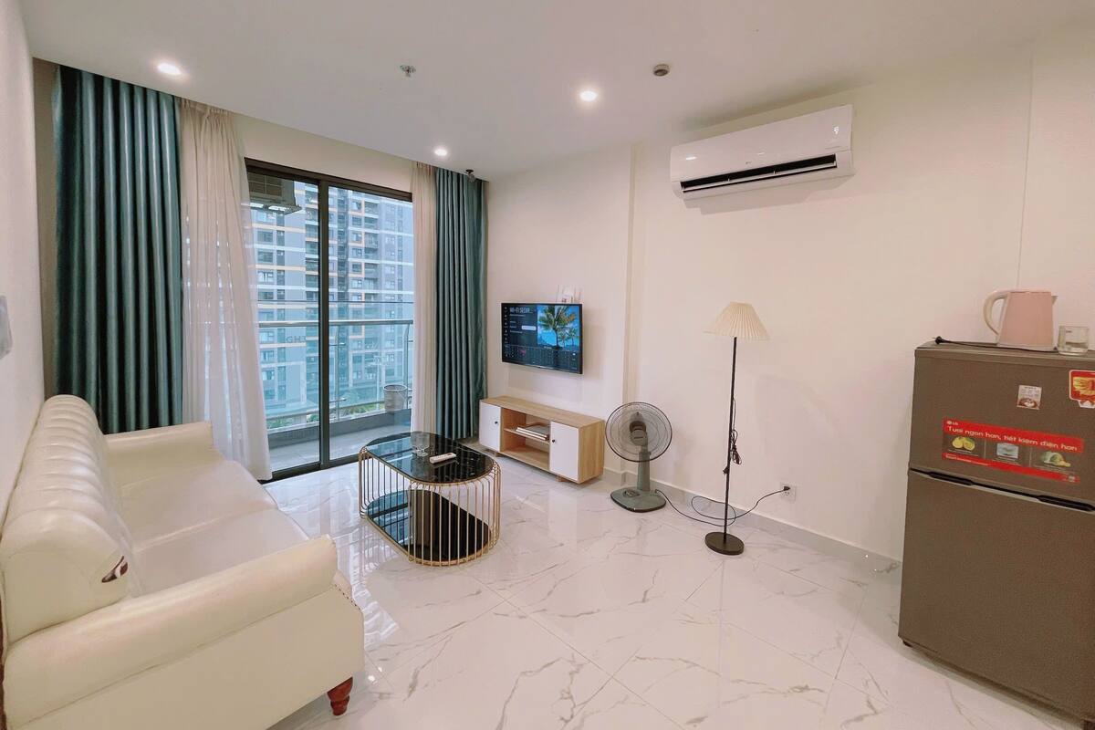 A modern living room with a sofa, a TV, an air conditioner, a refrigerator, and a view of high-rise buildings.