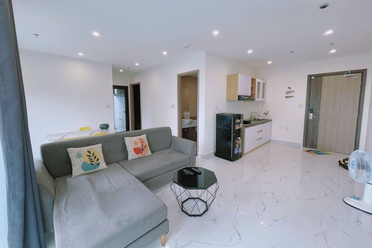 A modern living room with a grey sectional sofa, a small black coffee table, and a kitchen area with white cabinets and a black refrigerator.