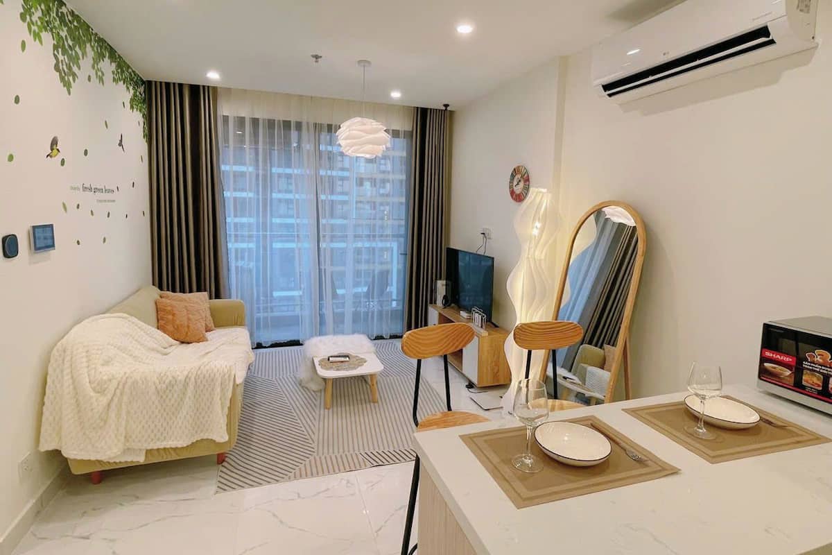 A modern hotel room with a sofa, a small table, a mirror, and a dining area with a glass of wine.