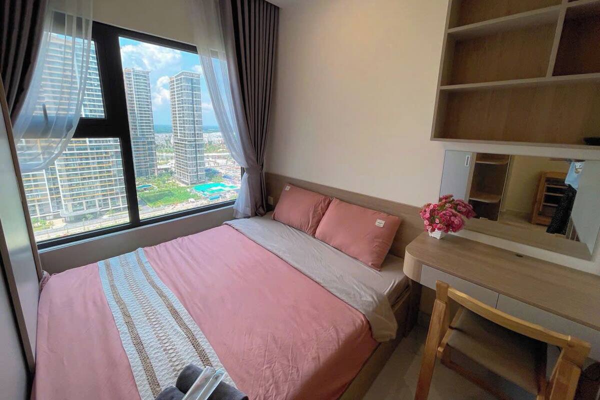 A hotel room with a large bed, a desk, a mirror, and a view of tall buildings through the window.