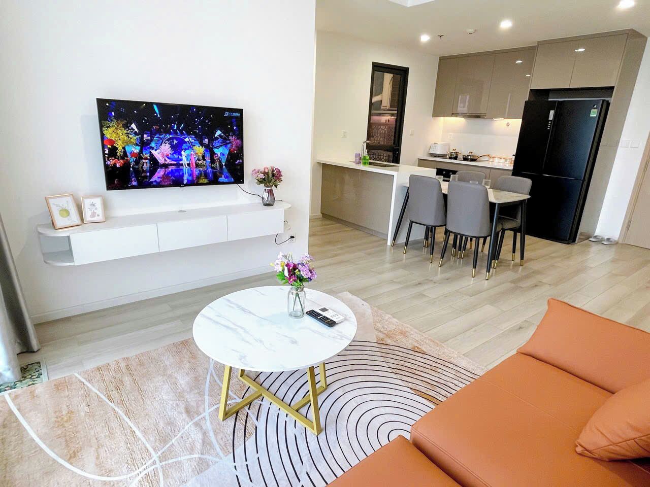 A modern living room with a flat-screen TV, a round coffee table, and a sofa. There is a kitchen area with a refrigerator and a dining table with chairs.