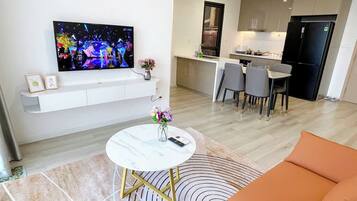 A modern living room with a flat-screen TV, a round coffee table, and a sofa. There is a kitchen area with a refrigerator and a dining table with chairs.