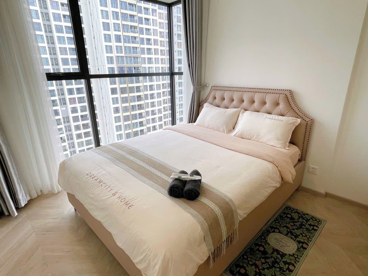 A bedroom with a large window, a bed with a beige headboard, and a pair of rolled-up towels on the bed.