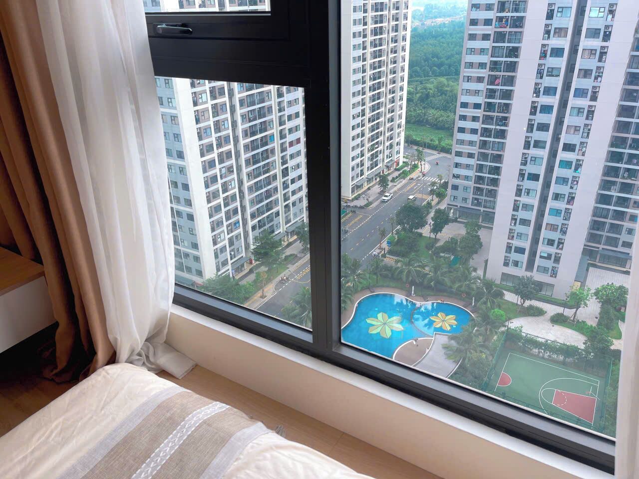 A bedroom with a large window overlooking a residential area with high-rise buildings and a swimming pool.