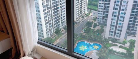 A bedroom with a large window overlooking a residential area with high-rise buildings and a swimming pool.