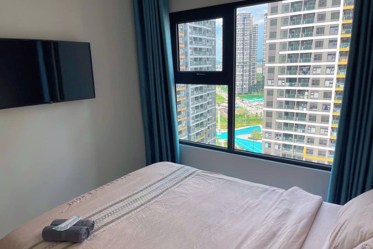A hotel room with a bed, a flat-screen TV mounted on the wall, and a window with a view of high-rise buildings and a pool.