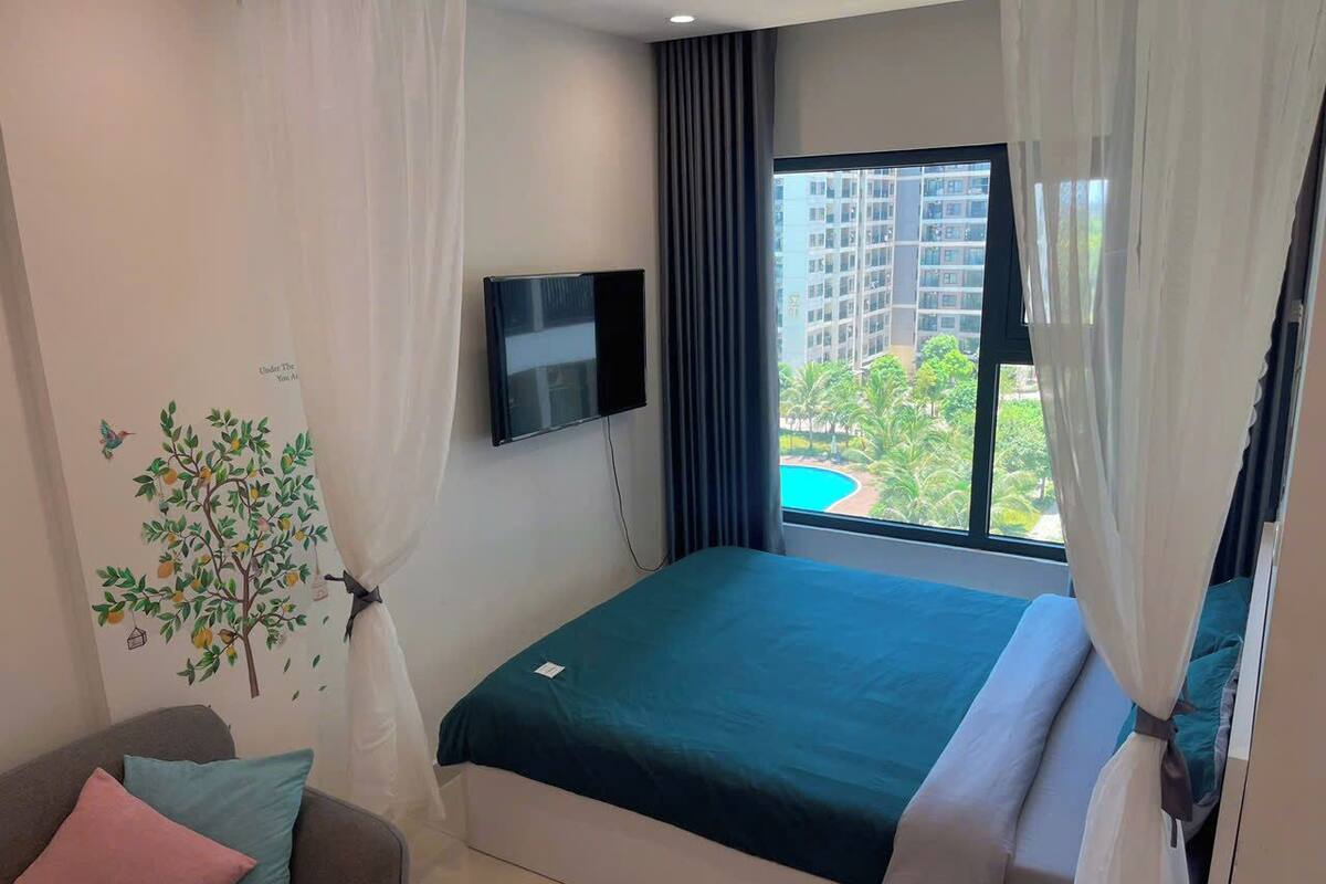 A modern hotel room with a large bed, a flat-screen TV, a window with a view of buildings and greenery, and a wall decal of a tree.