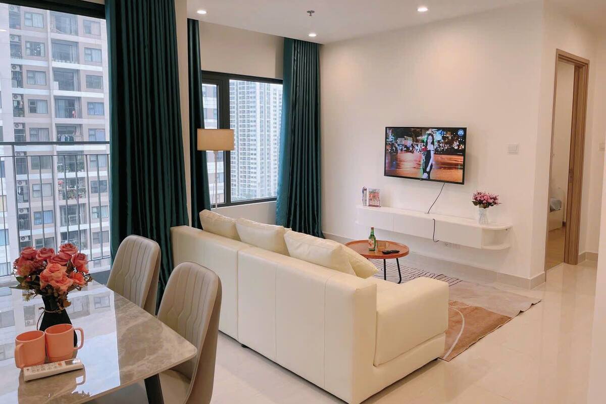 A modern living room with a white sofa, a dining table with chairs, a TV mounted on the wall, and a large window with curtains.