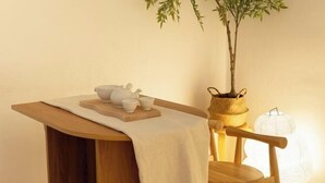 Una mesa de madera con un mantel blanco, una tetera, tazas y una pequeña planta en una cesta tejida.