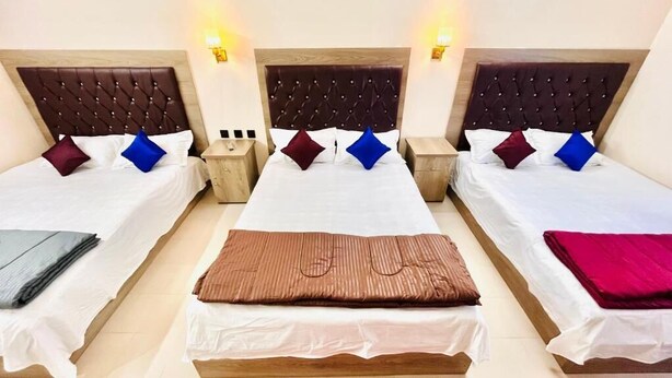 A hotel room with three beds, each with a wooden headboard and white bedding. There are two bedside tables with lamps and a small table between the beds. - Hotel Travellers Inn Sylhet (Sylhet)
