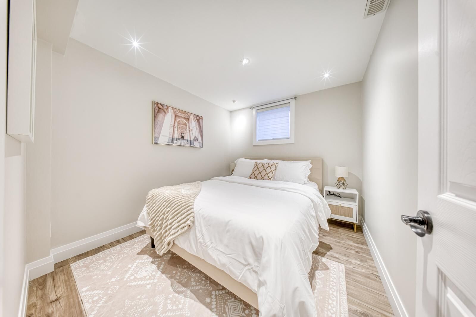 Une chambre à coucher avec un grand lit, une table de chevet, une commode, une fenêtre avec des stores et un luminaire au plafond.