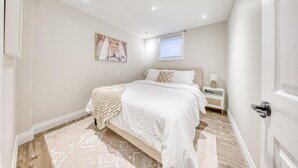 A bedroom with a large bed, a bedside table, a nightstand, a window with blinds, and a ceiling light. - The Cozy Cove on Flatford (Mississauga)