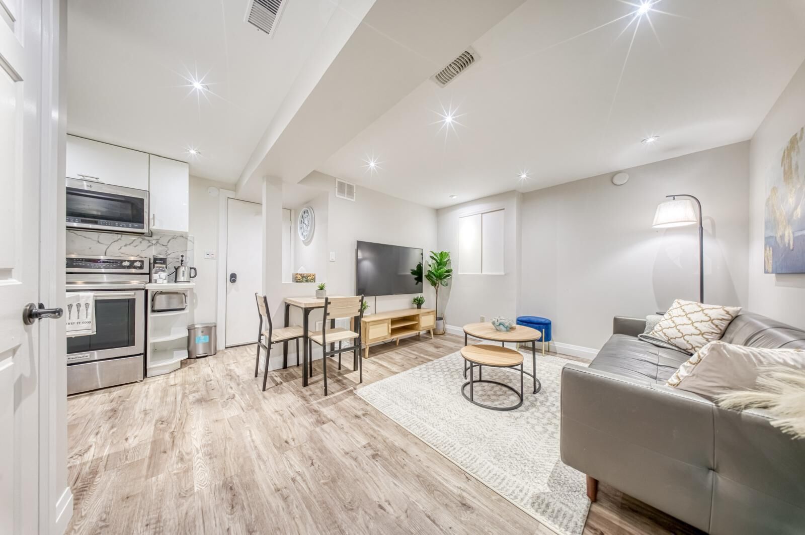 A modern living room with a grey sofa, a coffee table, and a TV. There is a kitchen area with a microwave, oven, and cabinets. The room has wooden flooring and a large window.